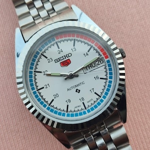 May include: A silver-toned Seiko 5 automatic wristwatch with a white face, featuring a day-date display and a blue and red accented dial. The watch has a stainless steel band and is marked "SEIKO 5 AUTOMATIC JAPAN MADE".