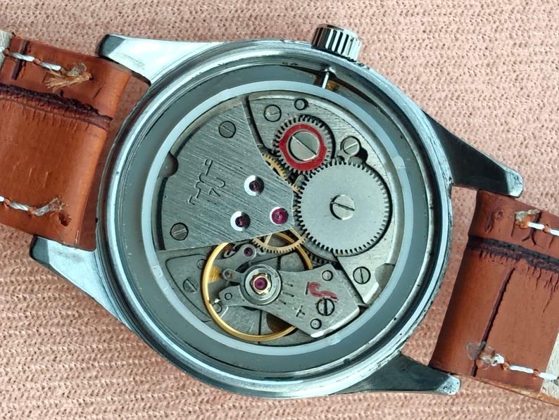 May include: Close-up of a vintage wristwatch with a silver case and a brown leather strap. The watch's intricate mechanical movement is visible, featuring gears, jewels, and a balance wheel. The watch has a textured silver crown.