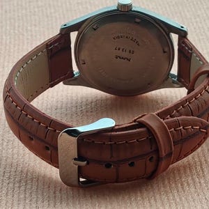 May include: A vintage wristwatch with a brown leather band and a silver-colored metal case. The band is stitched with a contrasting thread. The watch face is not visible. The watch is on a light brown surface.