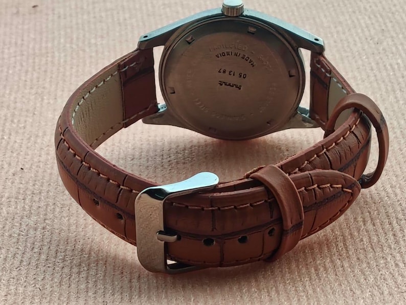 May include: A vintage wristwatch with a brown leather band and a silver-colored metal case. The band is stitched with a contrasting thread. The watch face is not visible. The watch is on a light brown surface.