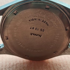 May include: Close-up of the back of a wristwatch with a stainless steel case and a brown leather strap. The watch face has text that reads "PROTECTED MADE IN INDIA" and "STAINLESS STEEL". The watch also has the numbers "05 13 87".
