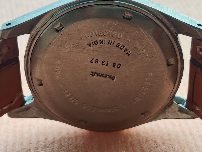 May include: Close-up of the back of a wristwatch with a stainless steel case and a brown leather strap. The watch face has text that reads "PROTECTED MADE IN INDIA" and "STAINLESS STEEL". The watch also has the numbers "05 13 87".