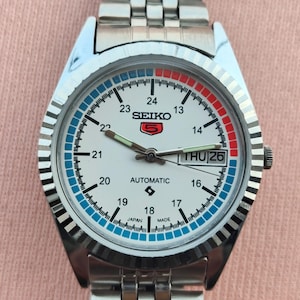 May include: A silver-toned Seiko automatic wristwatch with a white face, featuring blue and red accents. The watch has a day-date display and a stainless steel band. The watch face displays the text "SEIKO" and "AUTOMATIC".