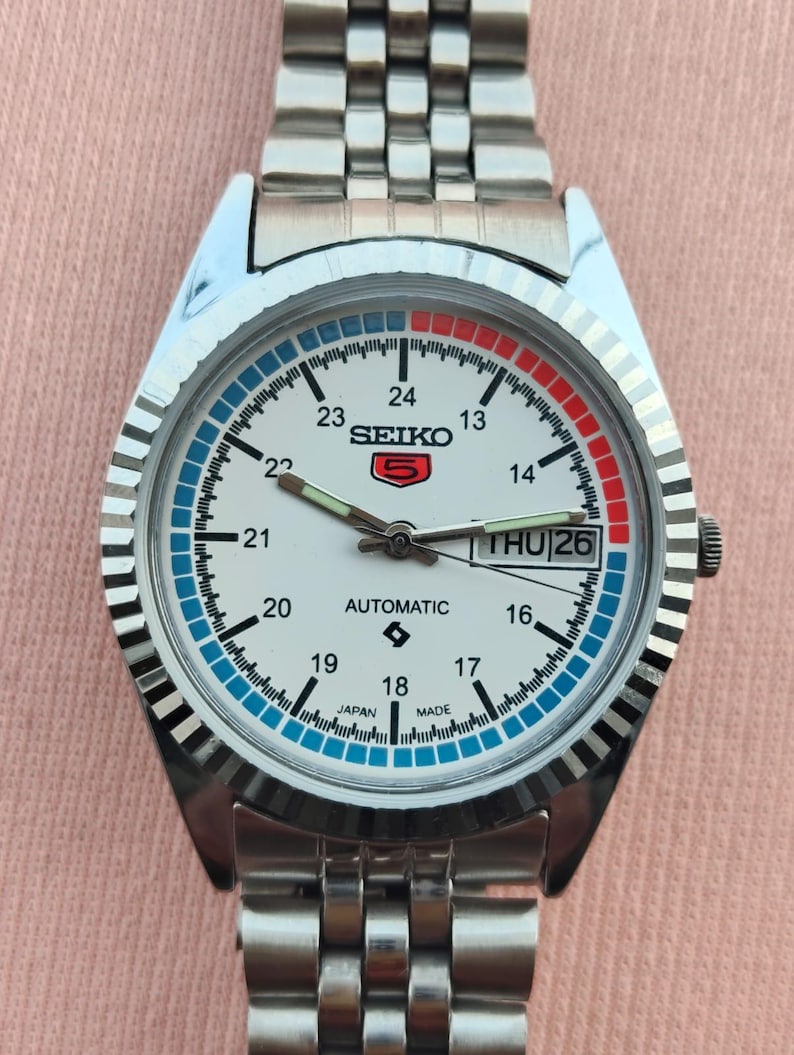 May include: A silver-toned Seiko automatic wristwatch with a white face, featuring blue and red accents. The watch has a day-date display and a stainless steel band. The watch face displays the text "SEIKO" and "AUTOMATIC".