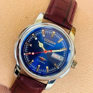 May include: A Citizen automatic wristwatch with a blue dial and a brown leather strap. The watch face displays the day and date, with the text "CITIZEN AUTOMATIC" and "21 JEWELS". The watch has a silver-colored case and gold-colored hands.