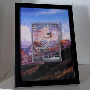 Op de afbeelding: Een ingelijste Charizard EX Pokémon-ruilkaart. De kaart zit in een beschermhoes, tegen een landschapsachtergrond. Het zwarte frame omringt de kaart, met een rode en oranje draak.