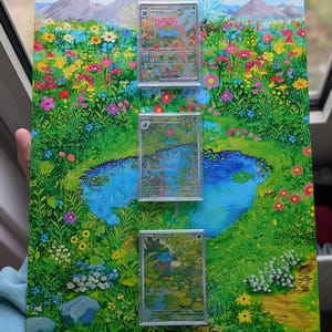 Könnte beinhalten: Ein rechteckiges Display-Board mit einer lebendigen, farbenfrohen Illustration einer üppigen Wiese mit einem zentralen blauen Teich, Bergen und Blumen. Drei durchsichtige Kunststoffhüllen mit Sammelkarten sind vertikal auf der Oberfläche montiert.