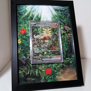 Könnte beinhalten: Gerahmte Pokémon-Karte mit "Gouging Fire ex" vor einem Dschungel-Hintergrund. Die Karte ist in einer transparenten Schutzhülle eingeschlossen und vor einem lebendigen grün-roten Blumendruck platziert. Der Rahmen ist schwarz.