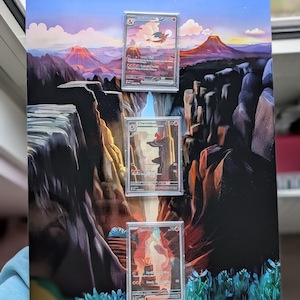 Può includere: Un pannello espositivo rettangolare con un paesaggio stampato di un canyon al tramonto e tre custodie in plastica trasparente montate verticalmente. Le carte all'interno sono etichettate come Charizard, Charmeleon e Charmander.