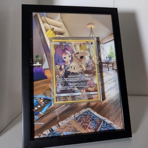 Op de afbeelding: Een ingelijste Pokémon-ruilkaart met Mimikyu, met een karakterillustratie en tekst. De kaart wordt weergegeven in een zwart frame, tegen een gedetailleerde achtergrondafbeelding van een kamer met een tapijt en naaispullen.