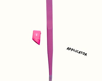 Puede incluir: Una herramienta aplicadora rosa con una punta y una pequeña tapa rosa separada. La palabra "APPLICATOR" está escrita en texto negro en ángulo.