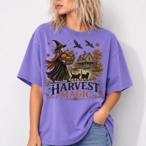 Puede incluir: Una camiseta morada con un estampado gráfico de una bruja con sombrero que lleva una cesta de calabazas, dos gatos negros, una casa y pájaros. El texto HARVEST MAGIC está impreso en negro y naranja debajo de la ilustración.