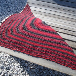 May include: A handmade, triangular shawl in red and black stripes. The shawl is knitted with a detailed pattern and has a scalloped edge. It is displayed on a wooden bench, showcasing its texture and design.