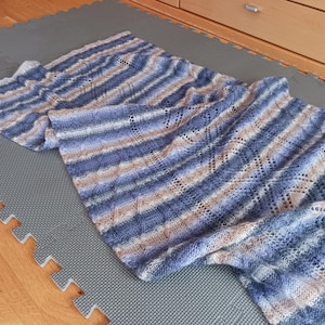 May include: A blue, tan, and white striped knitted scarf. The scarf is laid out on a gray surface. The scarf has a delicate, openwork design and is made of soft yarn. The scarf is a rectangular shape.