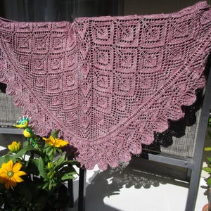 Puede incluir: Un chal tejido de color malva con un patrón cuadrado y bordes festoneados está drapeado sobre una silla. El intrincado diseño del chal es visible, mostrando la detallada artesanía. El chal tiene forma triangular, perfecto para usar.