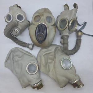 Może przedstawiać: Kolekcja pięciu zabytkowych masek gazowych w różnych stylach. Maski są głównie szare z przezroczystymi soczewkami i dołączonymi wężami lub filtrami. Niektóre maski mają konstrukcję z pełnym kapturem, inne bardziej tradycyjną maskę na twarz.