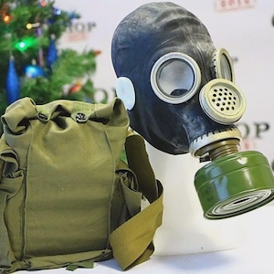 Op de afbeelding: Een zwart gasmasker met ronde oogstukken en een groen filter wordt getoond naast een groene canvas tas. Het masker staat op een witte standaard. De tas heeft meerdere zakken en een schouderriem.
