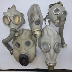 Puede incluir: Cinco máscaras de gas vintage en varios estilos y condiciones. Las máscaras son principalmente grises con oculares transparentes y algunas tienen mangueras adjuntas. Las máscaras están dispuestas sobre una superficie blanca.