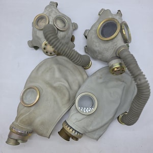 Op de afbeelding: Vier vintage gasmaskers in lichtgrijze kleur. Elk masker heeft een cirkelvormige lens, een gegolfde slang en een schroefdraadconnector. De maskers lijken gemaakt van rubber en metaal.