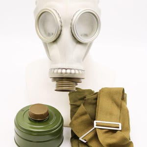 Könnte beinhalten: Eine Vintage-Gasmaske mit weißem Gummi-Gesichtsteil, klaren kreisförmigen Augenlinsen und einem Gewindeanschluss. Enthält einen grünen Filterbehälter und eine khakifarbene Canvas-Tasche.