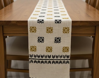 Op de afbeelding: Een crèmekleurige tafelloper met een herhalend geometrisch patroon in zwart en goud. De loper is aan de uiteinden gefranjerd en wordt gepresenteerd op een houten tafel. De tafelloper is ongeveer 30 cm breed.