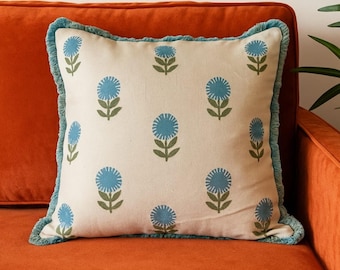 May include: Square throw pillow with a cream-colored background and a repeating pattern of blue floral designs. The pillow has a blue fringe trim and is placed on an orange sofa. The floral pattern includes blue flowers and green stems.