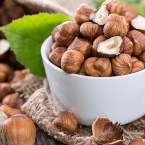 May include: A white ceramic bowl filled with shelled brown hazelnuts. The nuts have a round, textured appearance with some showing a split white interior. A single green leaf and pieces of burlap fabric are visible near the bowl of nuts.