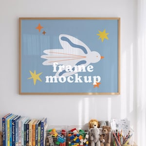 Può includere: Un mockup di stampa incorniciata con uno sfondo azzurro chiaro con un disegno di uccello bianco e il testo "frame mockup". La cornice è in legno chiaro. Sotto, una mensola di legno contiene libri, giocattoli e materiale artistico.