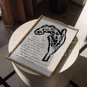 May include: A framed art print featuring a black and white abstract floral design with handwritten French text. The artwork is displayed on a white table, with a neutral-toned rug and curtains in the background. The frame is a light wood color.