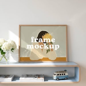 May include: A framed art print mockup with the text "frame mockup" in white. The artwork features a person holding a yellow flower. A blue and white toy van sits on a shelf below the frame, with magazines and a vase of white flowers.