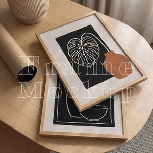 May include: Two framed art prints with minimalist botanical designs, featuring a monstera leaf and a vase, rest on a light wood table. A rolled-up poster and a potted plant are also visible. The frames have a natural wood finish.