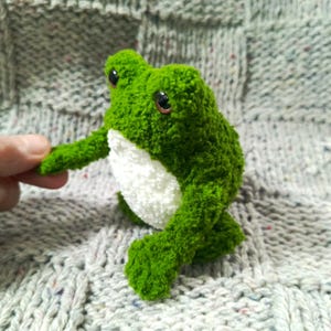 Puede incluir: Un peluche de rana verde hecho a mano con vientre blanco. La rana tiene ojos grandes y oscuros y está hecha de un material texturizado y difuso. El peluche está sobre una superficie de punto gris.
