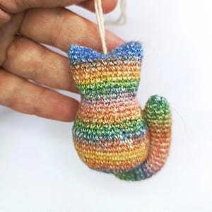 May include: A small, handmade cat ornament in a rainbow of colors, including blue, green, yellow, and orange. The cat is crocheted and has a small loop for hanging. The cat's tail is curled, and the ears are pointed.