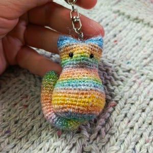May include: A small, crocheted cat keychain featuring a variegated yarn pattern in shades of orange, yellow, blue, green, and pink. The cat has two small black bead eyes, pointed ears, and a curved tail, attached to a silver-toned metal lobster clasp.