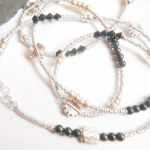 May include: Close-up of layered necklaces with silver beads, black beads, and decorative silver accents. The necklaces are arranged in a swirling pattern against a white background, creating a delicate and intricate design. The jewelry is suitable for everyday wear.