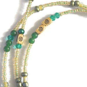 K&ouml;nnte beinhalten: Eine doppelreihige Perlenkette mit gr&uuml;nen und goldenen Perlen. Die Kette hat einen goldenen Anh&auml;nger mit einem dekorativen Muster.