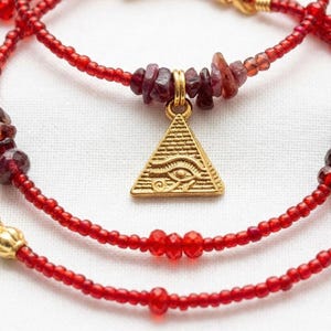 May include: A layered necklace featuring red seed beads and garnet accents. A gold-colored pyramid pendant with an eye design hangs from the center. Gold-colored beads are interspersed throughout the necklace.
