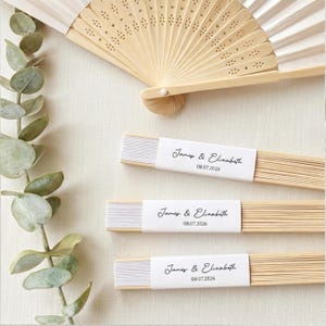 May include: A collection of wedding fans and eucalyptus leaves. The fans are made of light wood and white paper, with the names "James & Elisabeth" and the date "08.07.2026" printed on them. The fans are arranged on a light-colored surface.