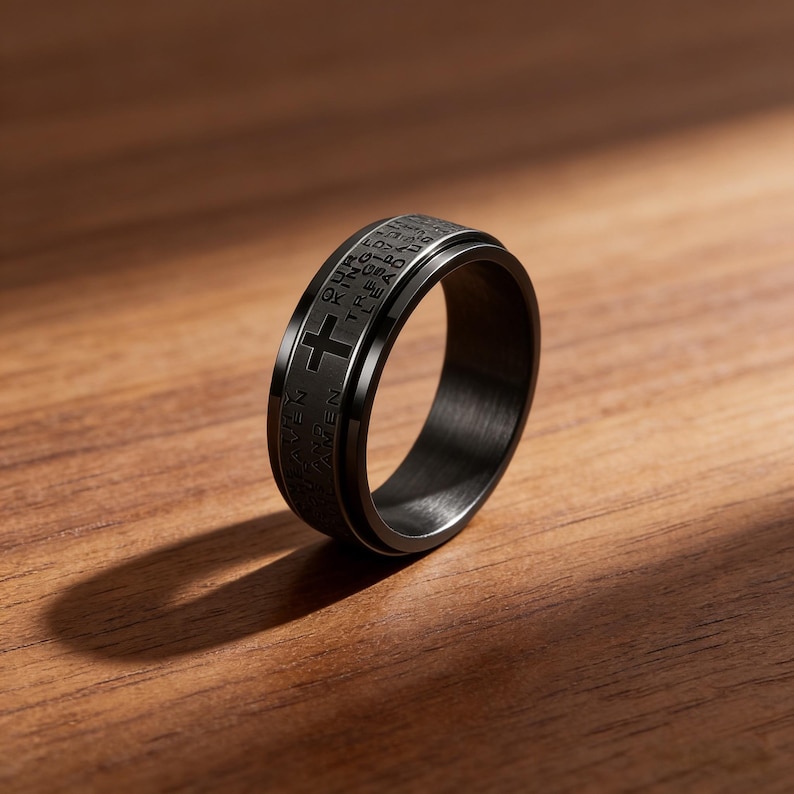 K&ouml;nnte beinhalten: Ein schwarzer Ring mit einem Kreuzdesign und eingraviertem Text. Der Ring besteht aus dunklem Metall und liegt auf einer Holzoberfl&auml;che. Das Design des Rings umfasst ein Kreuz und religi&ouml;sen Text.