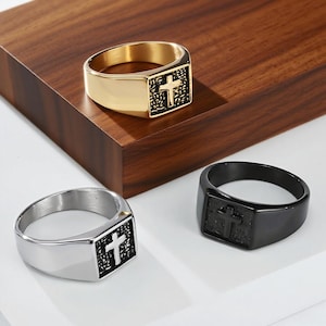May include: Three rings with a cross design. One ring is gold-colored, another is silver, and the third is black. Each ring features a rectangular face with a cross emblem. The rings are displayed on a wooden surface.
