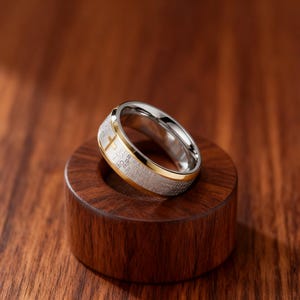 K&ouml;nnte beinhalten: Ein silber- und goldfarbener Ring mit Kreuzdesign. Der Ring hat ein goldenes Band und eine silberne Innenseite. Das &auml;u&szlig;ere Band hat eingravierten Text. Der Ring wird auf einem kleinen, dunklen Holzst&auml;nder pr&auml;sentiert.
