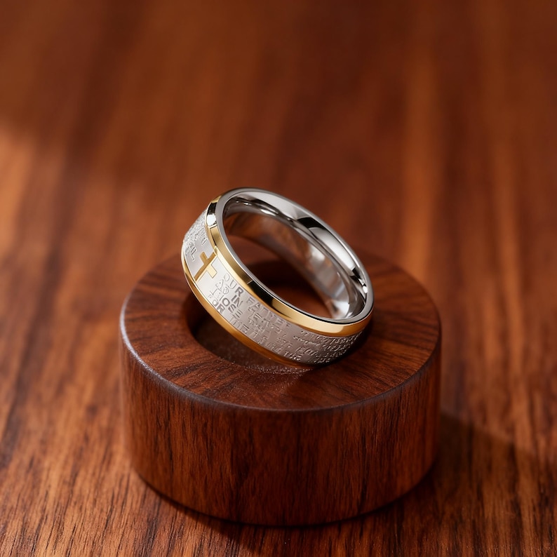 K&ouml;nnte beinhalten: Ein silber- und goldfarbener Ring mit Kreuzdesign. Der Ring hat ein goldenes Band und eine silberne Innenseite. Das &auml;u&szlig;ere Band hat eingravierten Text. Der Ring wird auf einem kleinen, dunklen Holzst&auml;nder pr&auml;sentiert.