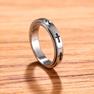 May include: A silver-colored ring with a series of small, black cross designs etched around the band. The ring is set against a wooden surface, highlighting its metallic sheen. The ring's design is simple and elegant, suitable for everyday wear.