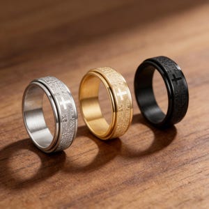K&ouml;nnte beinhalten: Drei Ringe mit Kreuzdesign. Ein Ring ist silberfarben, einer goldfarben und einer schwarz. Jeder Ring hat ein Kreuz und Text um das Band. Die Ringe werden auf einer Holzoberfl&auml;che pr&auml;sentiert.