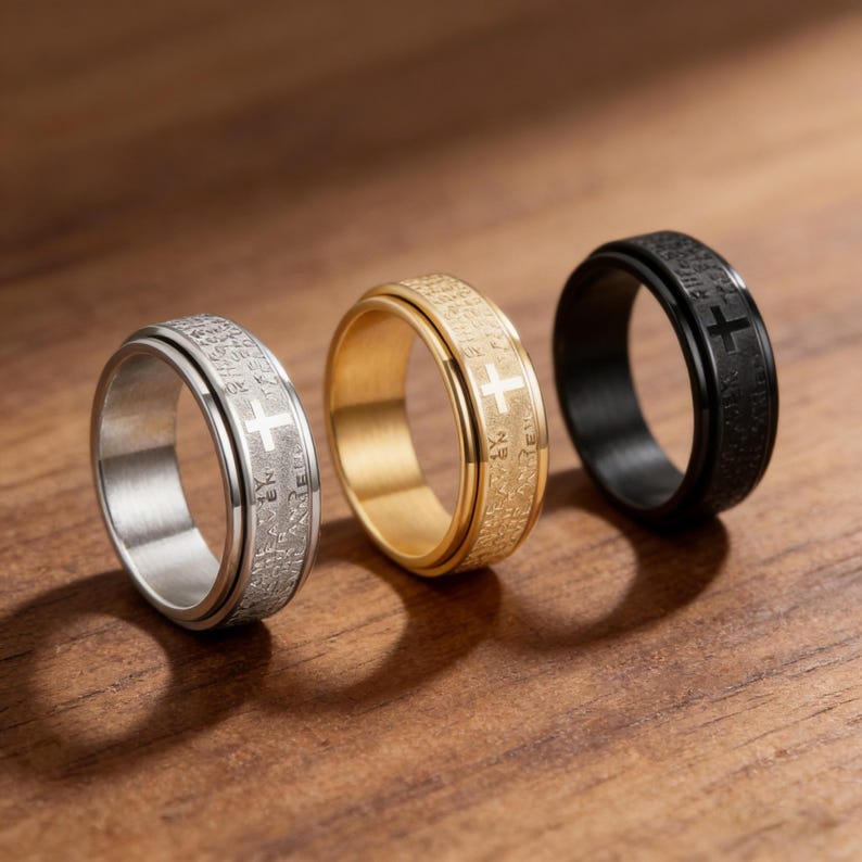 K&ouml;nnte beinhalten: Drei Ringe mit Kreuzdesign. Ein Ring ist silberfarben, einer goldfarben und einer schwarz. Jeder Ring hat ein Kreuz und Text um das Band. Die Ringe werden auf einer Holzoberfl&auml;che pr&auml;sentiert.