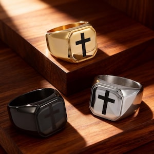 Peut inclure: Trois chevalières avec un motif de croix. Une bague est dorée, une argentée et une noire. Chaque bague présente une croix noire sur une face carrée. Les bagues sont présentées sur une surface en bois.