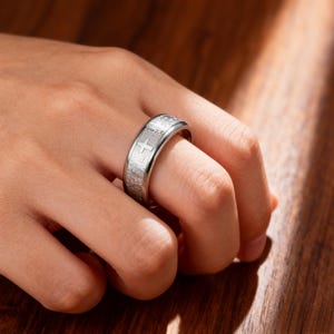 K&ouml;nnte beinhalten: Ein silberfarbener Ring mit Kreuzdesign und eingraviertem Text. Der Ring wird an einem Finger getragen und zeigt seine detaillierte Handwerkskunst. Die Oberfl&auml;che des Rings reflektiert das Licht und hebt die komplizierten Details des Designs hervor. Durchmesser ca. 2 cm.