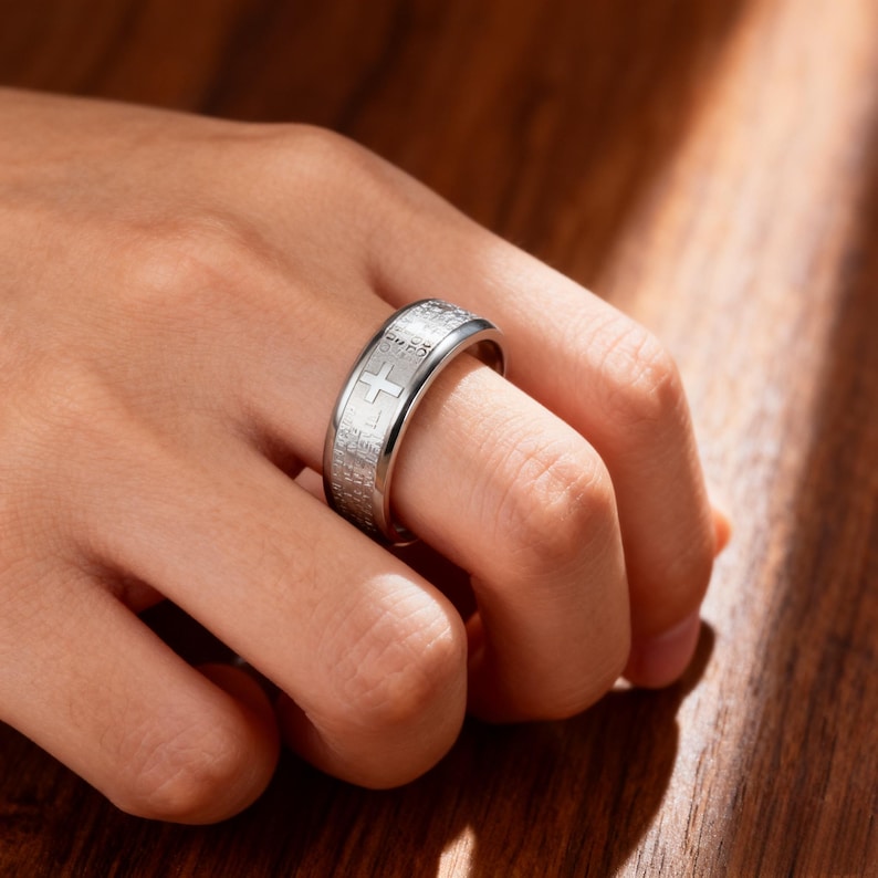 K&ouml;nnte beinhalten: Ein silberfarbener Ring mit Kreuzdesign und eingraviertem Text. Der Ring wird an einem Finger getragen und zeigt seine detaillierte Handwerkskunst. Die Oberfl&auml;che des Rings reflektiert das Licht und hebt die komplizierten Details des Designs hervor. Durchmesser ca. 2 cm.