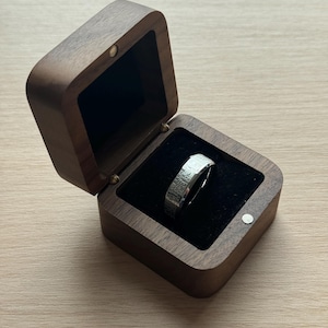 K&ouml;nnte beinhalten: Ein silberner Ring mit strukturierter Oberfl&auml;che liegt in einer offenen, quadratischen Holzbox mit schwarzem Samtinterieur. Die Box besteht aus dunkelbraunem Holz und hat einen Klappdeckel. Der Ring ist der Blickfang, pr&auml;sentiert auf dem schwarzen Samt.