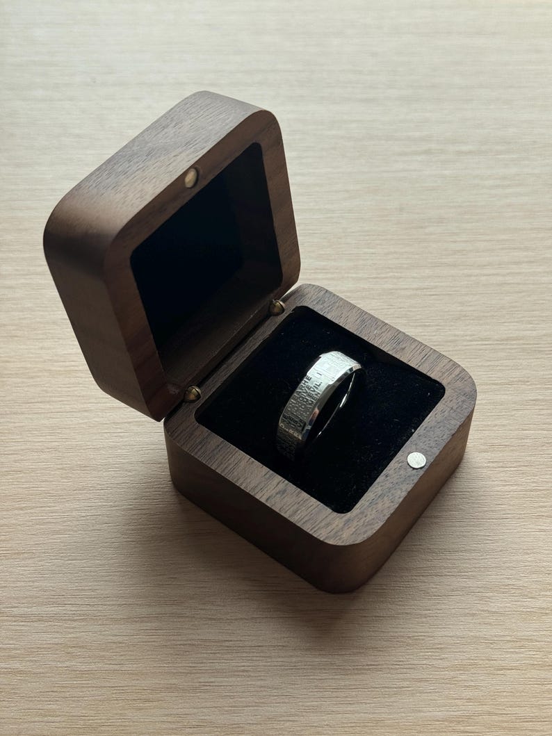 K&ouml;nnte beinhalten: Ein silberner Ring mit strukturierter Oberfl&auml;che liegt in einer offenen, quadratischen Holzbox mit schwarzem Samtinterieur. Die Box besteht aus dunkelbraunem Holz und hat einen Klappdeckel. Der Ring ist der Blickfang, pr&auml;sentiert auf dem schwarzen Samt.